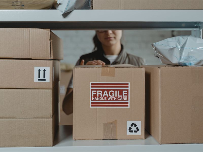 Colli pronti alla spedizione su una scaffalatura, con etichetta FRAGILE in primo piano e un&rsquo;operatrice al lavoro sullo sfondo